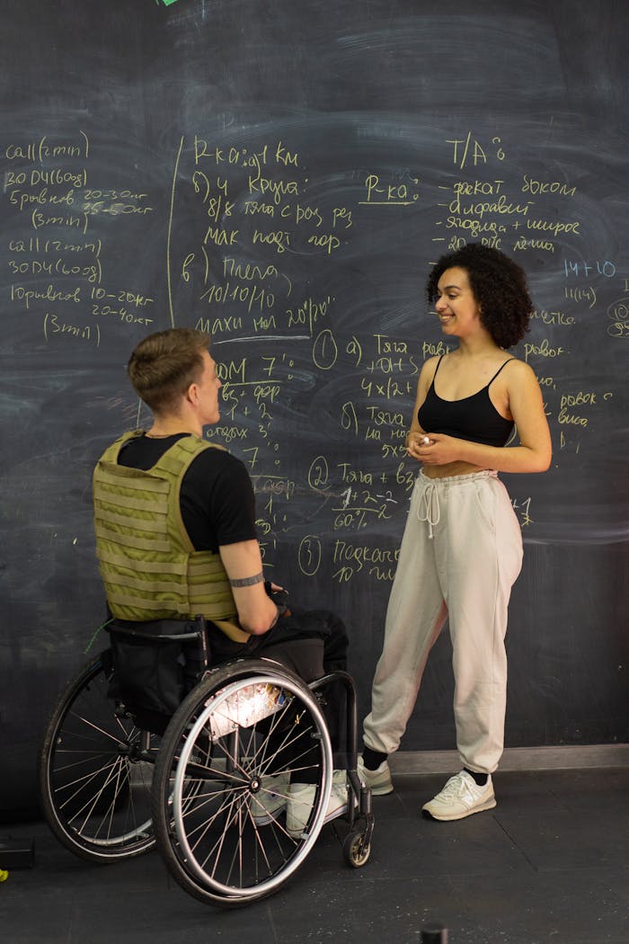 A female trainer and a disabled man in wheelchair discuss workout plans in gym setting.